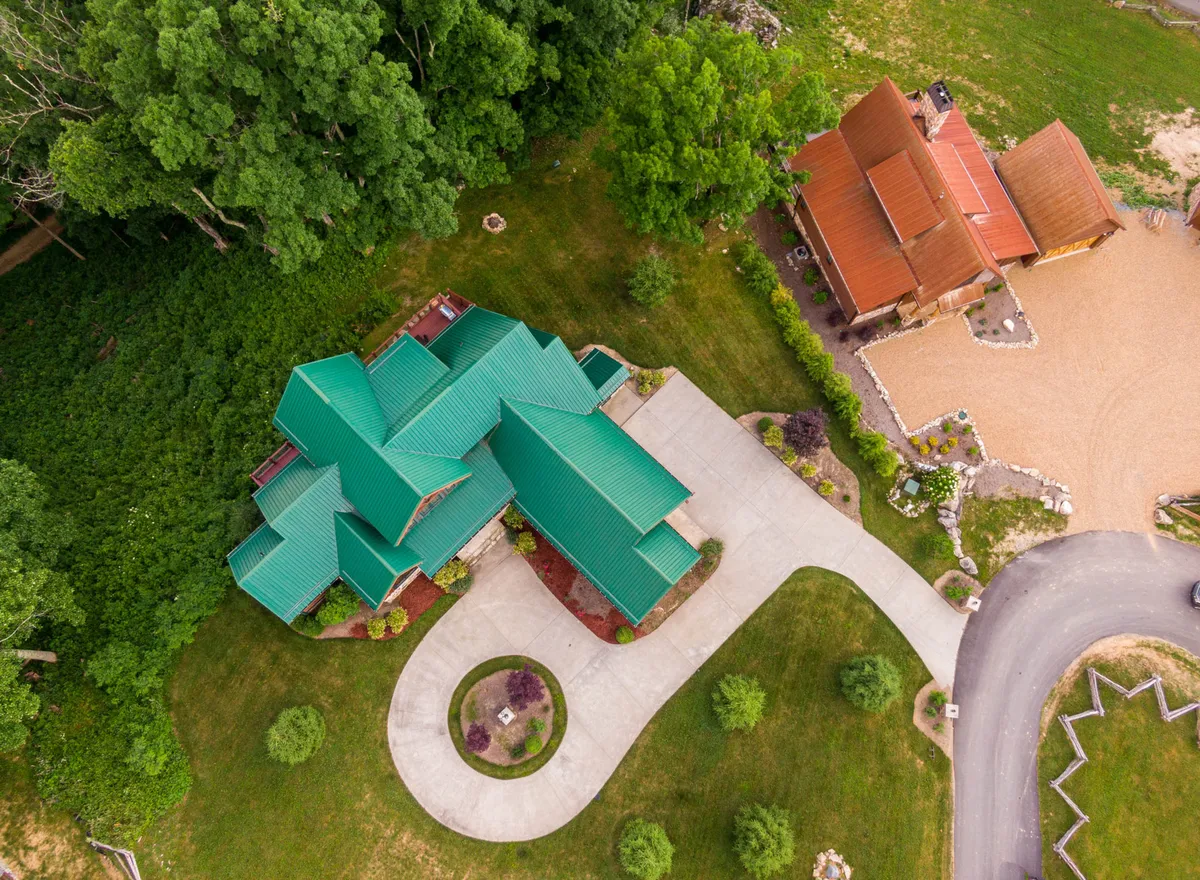 This image captures the house with a green roof, a curved driveway, and trees encircling the property. The presence of an adjacent building and the tranquil natural setting evoke opportunities for outdoor walks and exploration, aligning well with the pet-friendly and adventure-focused narrative.