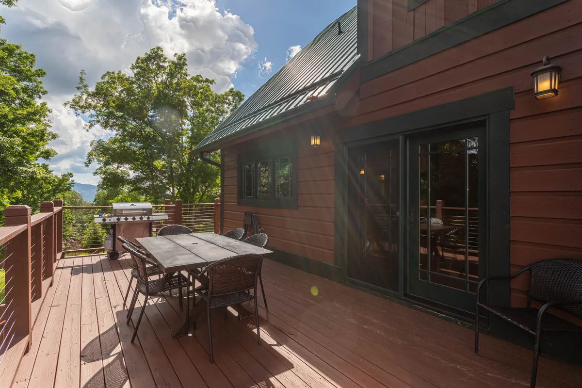 This image depicts a relaxed outdoor dining area on a deck surrounded by trees, featuring a dining table and a gas grill. The deck's railings and forested backdrop enhance the sense of privacy and immersion in nature, while the setup encourages gatherings, meals, and moments of connection outdoors.