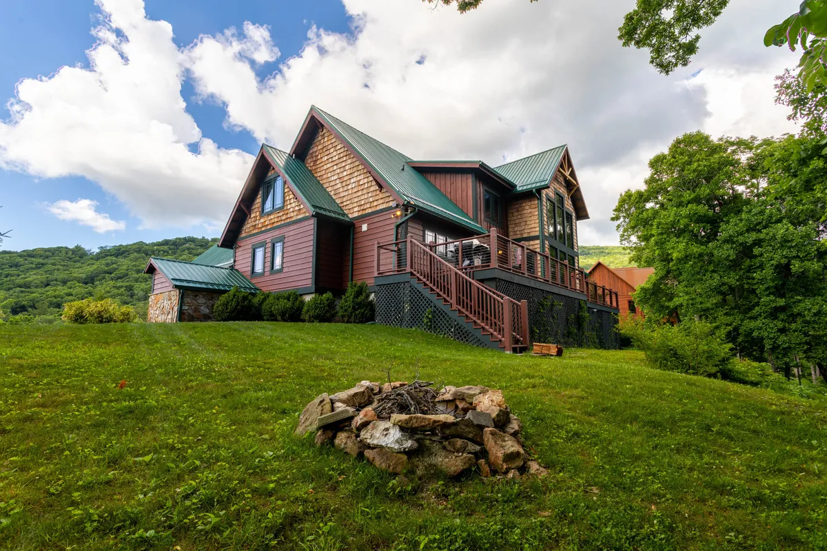 The image showcases the exterior of the house with a clear view of the stone fire pit area on a green lawn, inviting guests to gather for evenings by the fire. The house and surrounding natural landscape set a cozy, welcoming mood for outdoor gatherings and stargazing.
