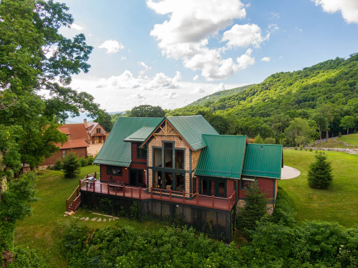 The exterior of the property features a striking green roof, large glass windows, and a spacious wooden deck, set against a serene natural backdrop. The scene hints at outdoor gatherings, relaxation, and easy access to nature.