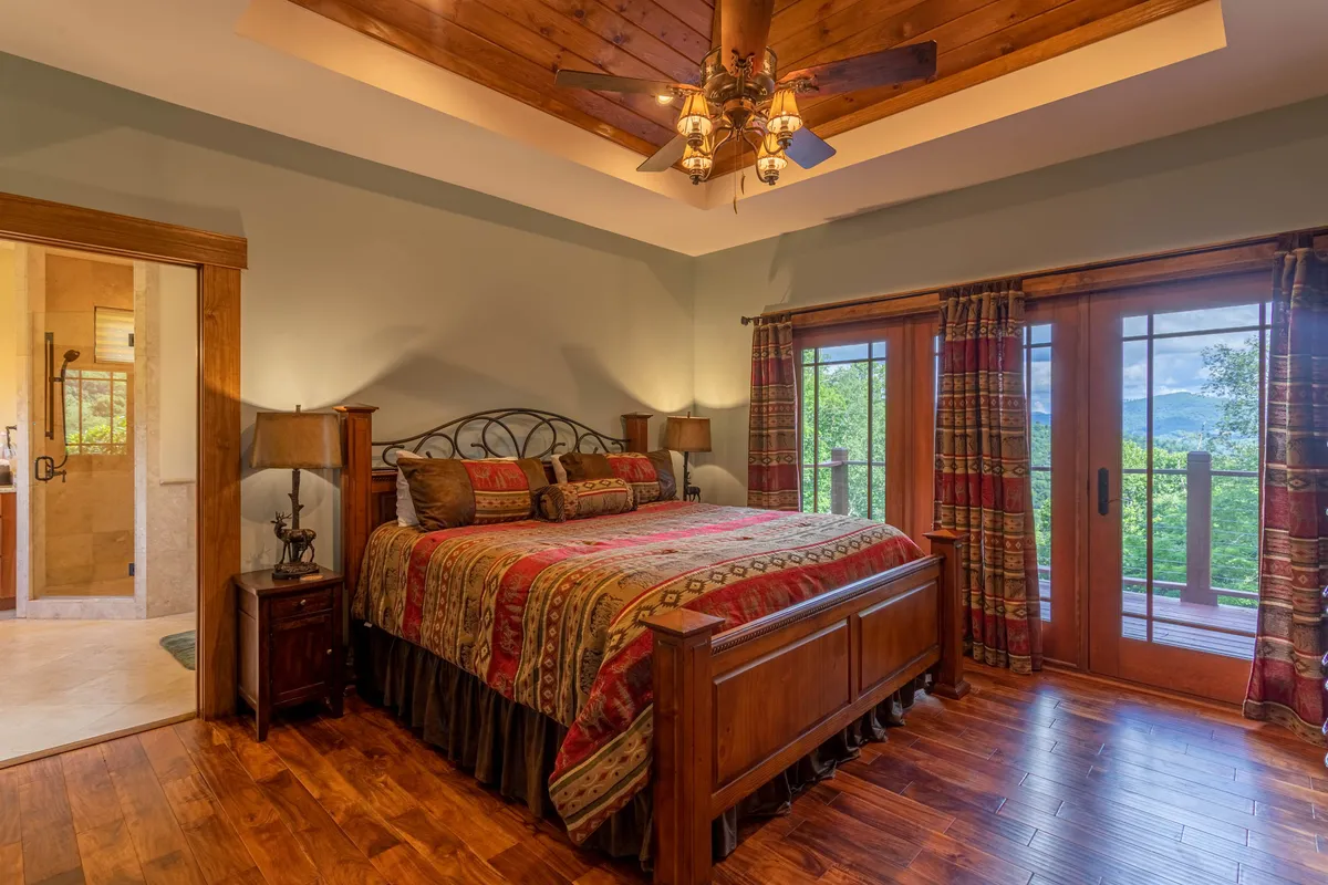 A queen bedroom featuring a comfortable bed with patterned bedding, a wooden ceiling fan overhead, and large windows letting in natural light and offering peaceful views. The room feels restful and inviting, ideal for relaxation.