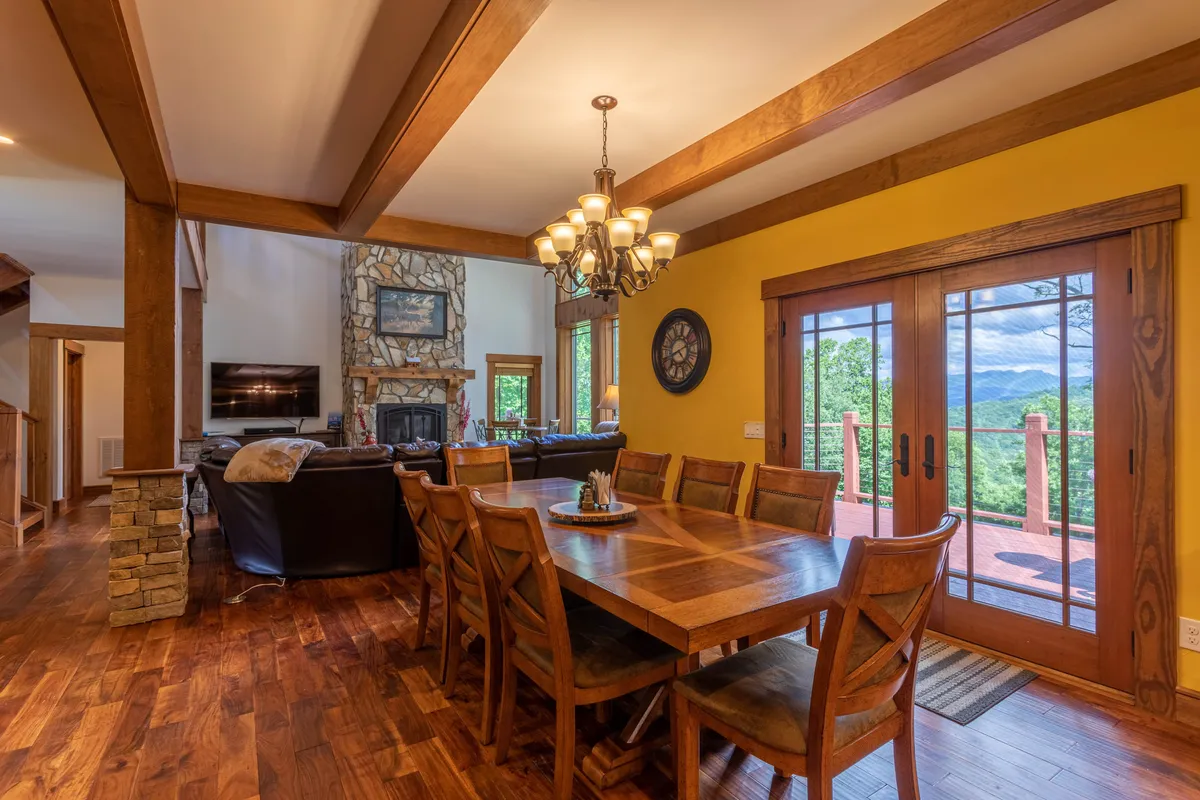 A warm and elegant dining area with a large wooden dining table set for eight, a beautiful chandelier overhead, a stone fireplace in the background, and large windows framing mountain views. The ambiance suggests shared meals and joyful conversations.