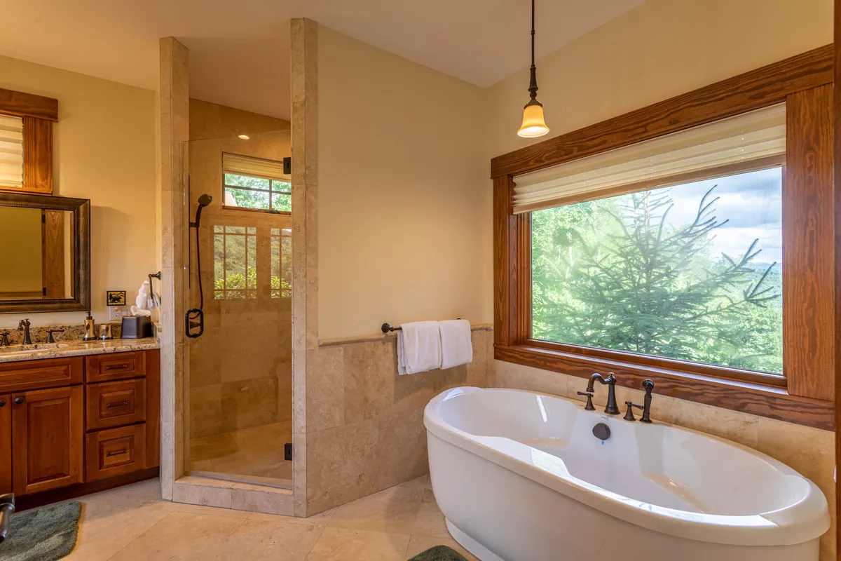 A serene, spa-inspired bathroom featuring a freestanding soaking tub positioned by a large window with tranquil nature views. The space boasts a walk-in tiled shower, a luxurious wooden vanity with a granite countertop, and an overall elegant, calming atmosphere.