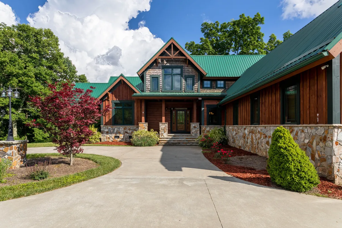 The image showcases the cabin's impressive stone facade, a charming green metal roof, and a welcoming front porch set among beautifully landscaped gardens. Rolling hills and natural greenery surround the home, while the porch offers a relaxing spot to take in stunning sunset views or enjoy a quiet morning coffee in nature.