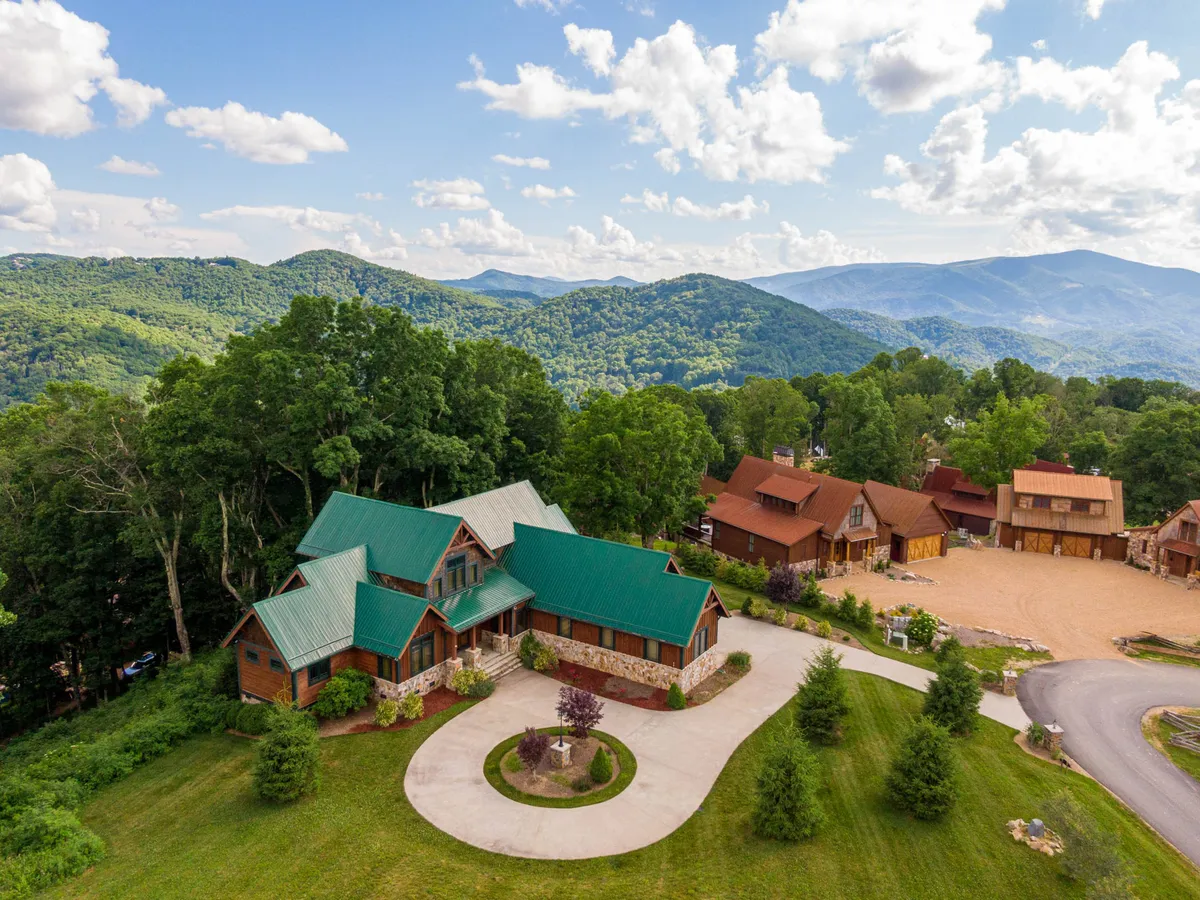 This image displays the main house with a green roof nestled among trees, with a stunning mountain backdrop and neighboring cabins visible. The setting suggests an immersive mountain experience and a direct connection to nature.