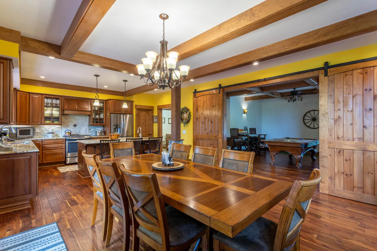 A welcoming interior space with a dining table, kitchen cabinetry, a striking chandelier, and prominent barn doors. The barn doors visually evoke the 'rustic sliding barn doors' mentioned in the section and suggest the flexible, expandable nature of the game room's connection to the main living area.