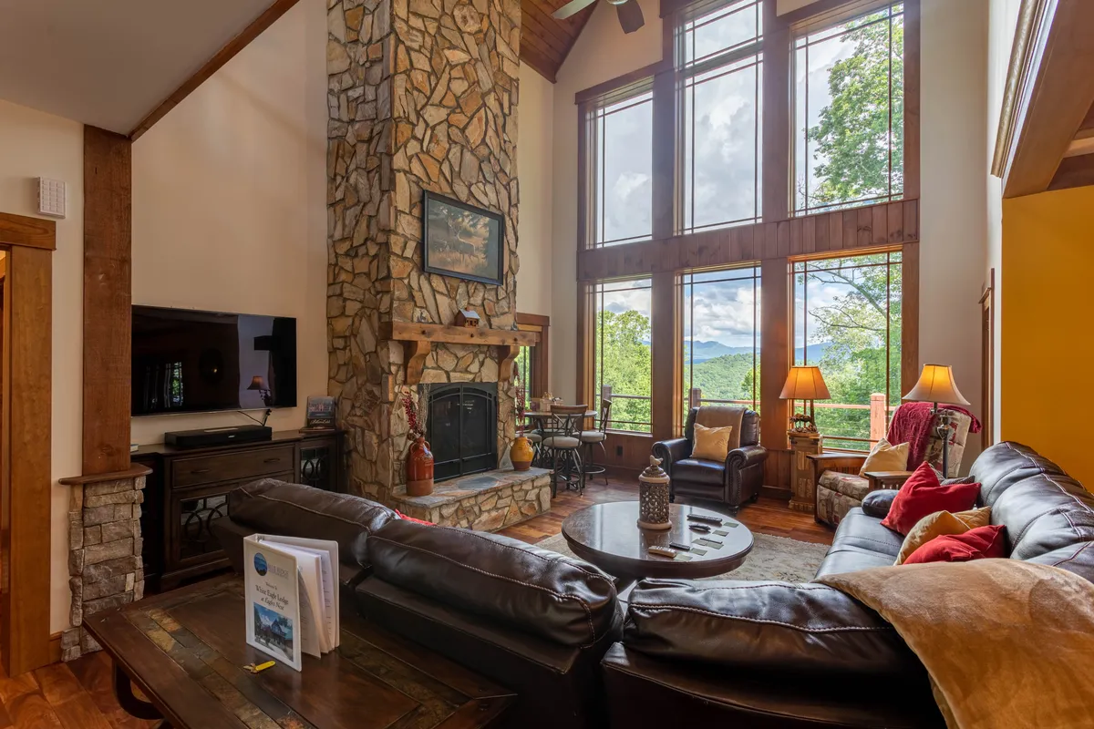 This image captures the open-concept living area featuring a grand stone fireplace, a plush leather sectional sofa, expansive windows revealing breathtaking mountain views, and a wooden coffee table set for cozy gatherings. The high ceilings and warm wood accents create a welcoming, spacious environment perfect for family and friends to gather.