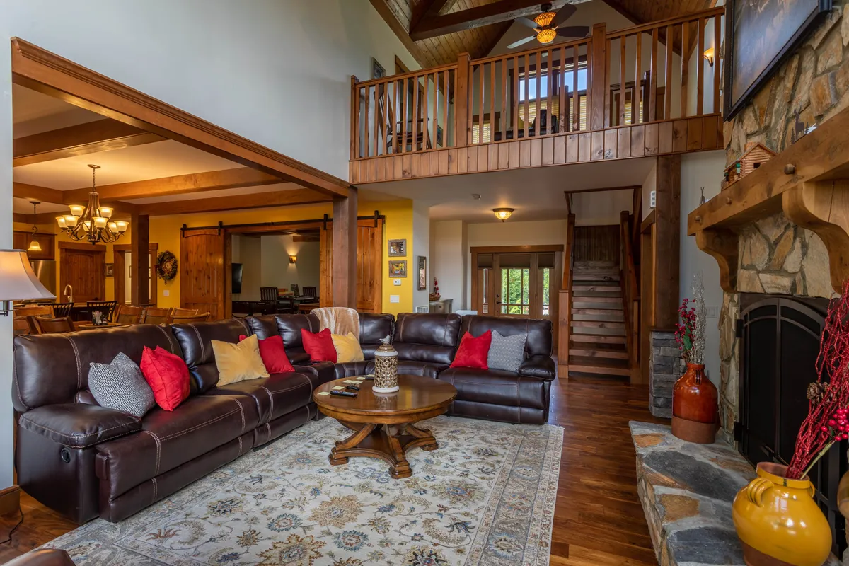 An inviting open living room with a large leather sectional sofa, round wooden coffee table, stone fireplace, and lofted ceilings with exposed wooden beams. The view also shows a staircase leading to the upper level, emphasizing the openness and interconnectedness of the space.