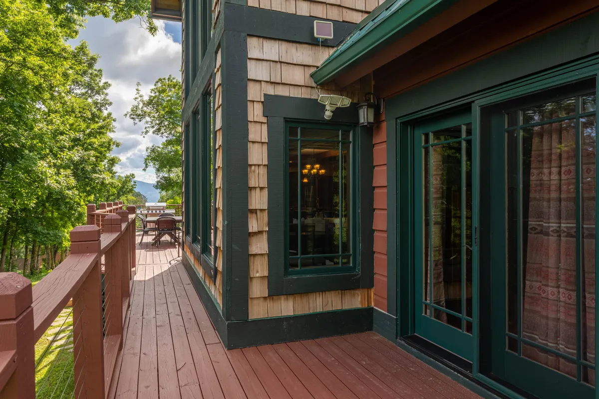 This image features a spacious deck with a sturdy railing, large windows that blur the lines between indoors and outdoors, wooden siding, and is surrounded by lush green trees. The vantage point emphasizes the seamless connection between the living space and the natural mountain landscape, inviting guests to relax and enjoy scenic views from the comfort of the deck.