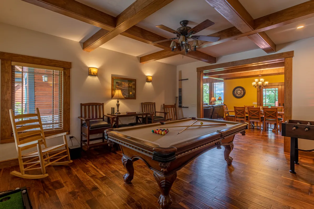 An inviting room centered around a pool table and accented by wooden beams, with a glimpse of a dining area and a cozy rocking chair. The setup conveys a casual, family-focused atmosphere where games and socializing blend seamlessly.